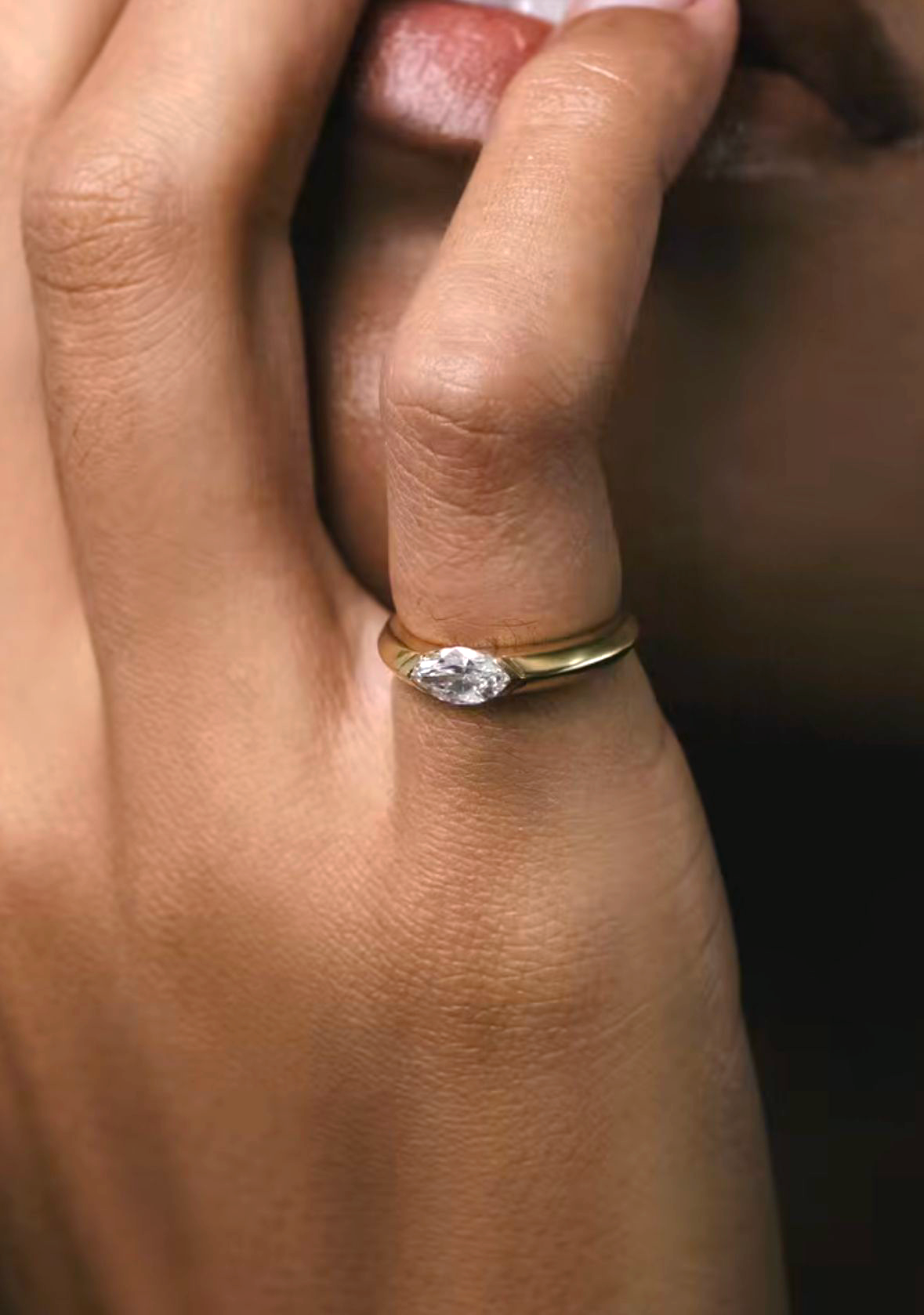 A bold yellow gold marquise ring worn on a finger, displaying an East-West oriented diamond in a sleek half-bezel setting. The substantial band design offers a modern, architectural look for a new heirloom.